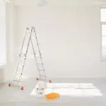 Bright, sunlit room prepped for interior painting with a ladder and supplies near large windows.