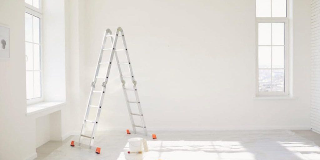 Bright, sunlit room prepped for interior painting with a ladder and supplies near large windows.